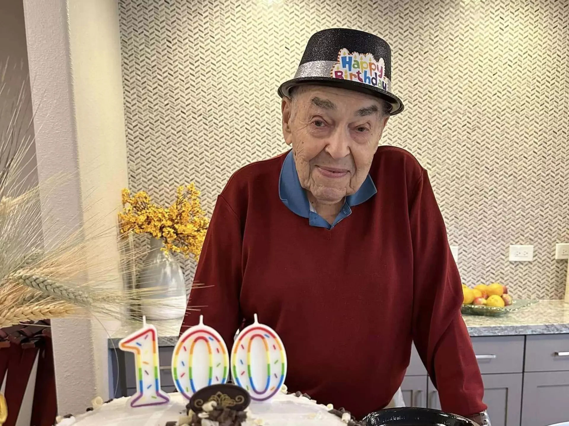 A centenarian poses in front of his cake, which has candles on it in the shape of the number 100.